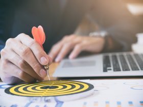 A person is holding a dart ready to aim at the bullseye of a target, with a laptop and business charts in the background, symbolizing goal setting and achievement in a business context.